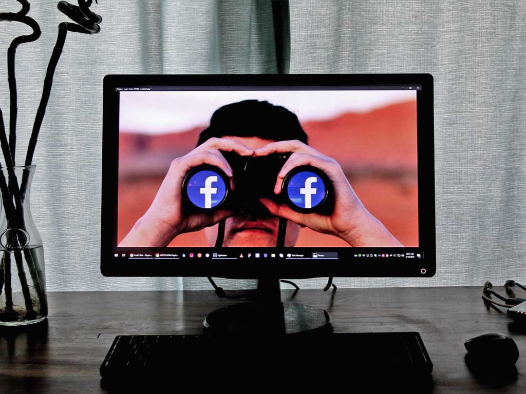 Shown on a computer monitor, a man with binoculars looks forward. Facebook logos are reflected in the lenses. 