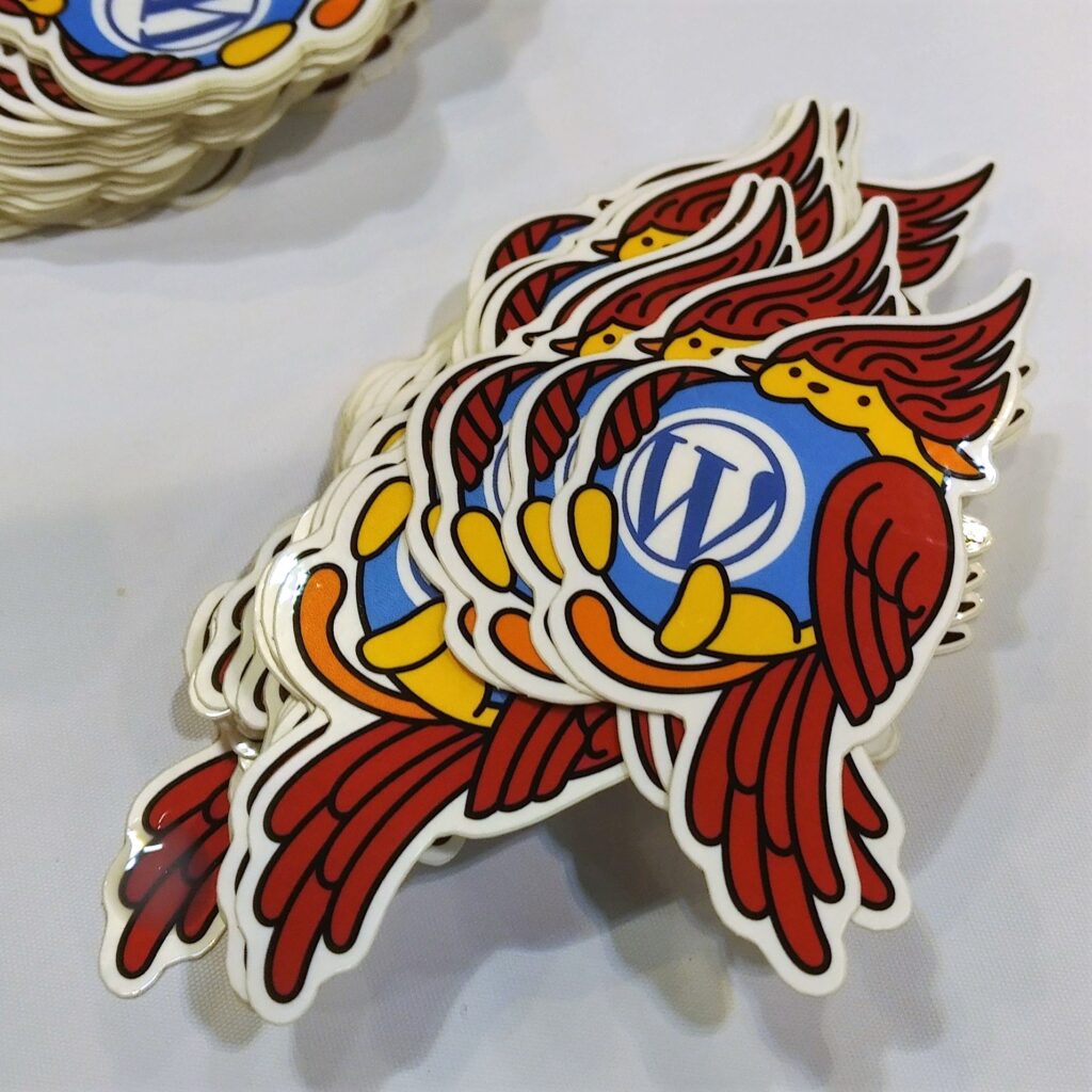 Stickers with Wapuu, the WordPress mascot, dressed as a cardinal, are shown on a table, honoring the host city, St. Louis. 